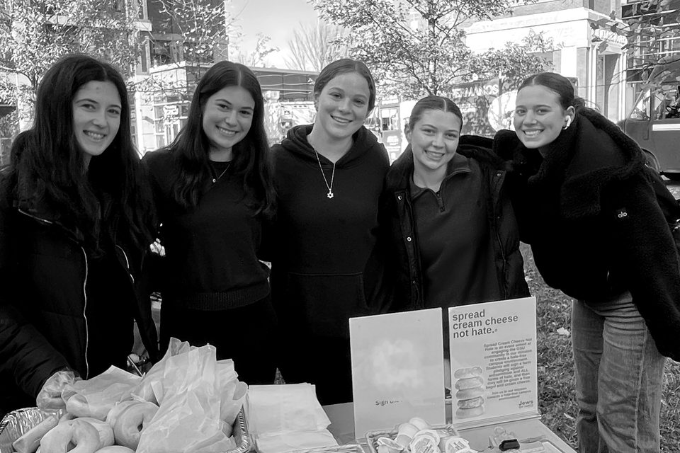 Five students stand behind a table of free bagels with a sign that reads, Spread cream cheese not hate.